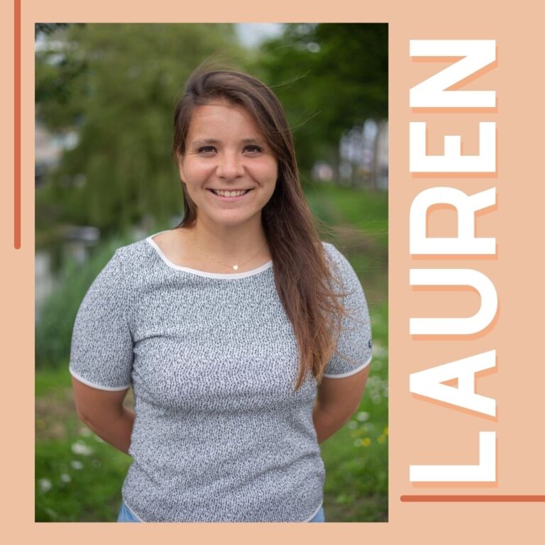 Vrouw met bruin stijl haar in een T-shirt. Ze heeft haar armen op haar rug en staat in een groene omgeving.